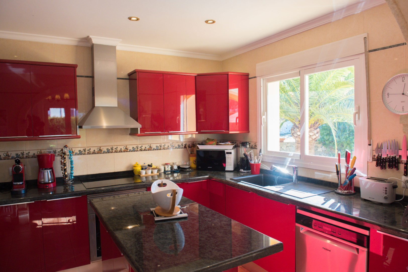 Cuisine équipée avec des façades rouges brillantes, des plans de travail en granit noir et une hotte en inox. La fenêtre apporte une lumière naturelle et vue sur le jardin.