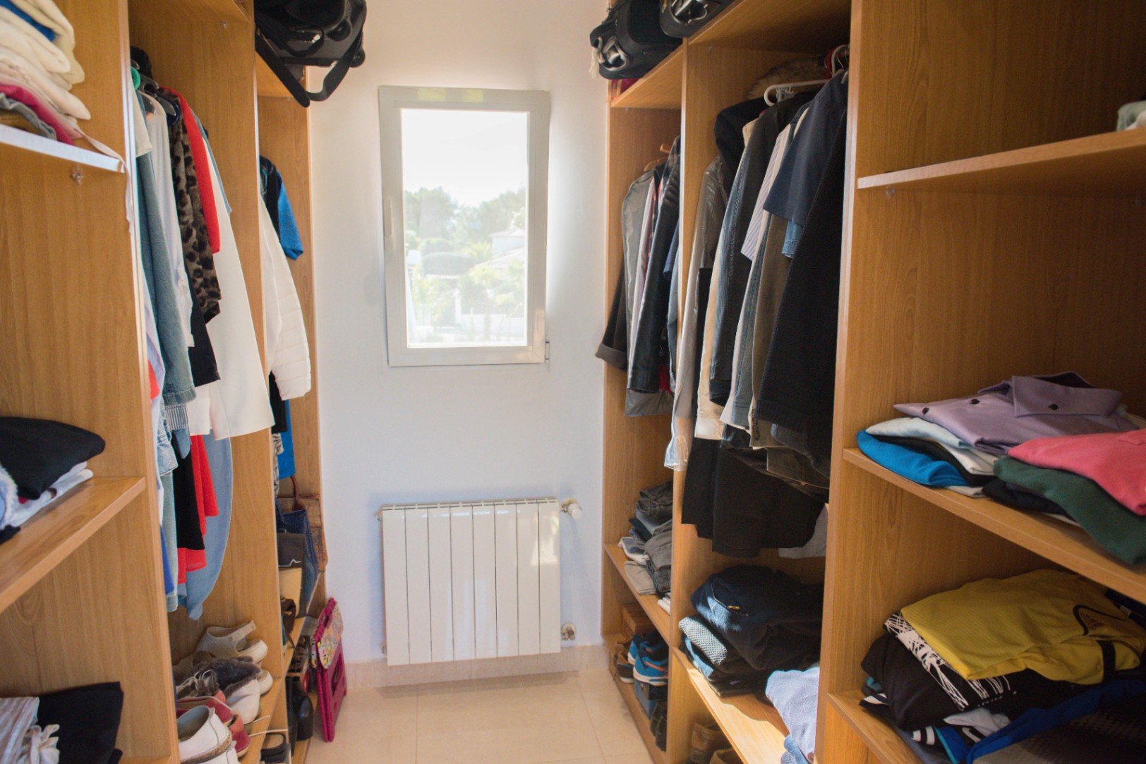 Dressing walk-in avec étagères en bois de chêne, penderies intégrées et rangements ouverts. Une fenêtre centrale apporte de la lumière naturelle au-dessus d'un radiateur.