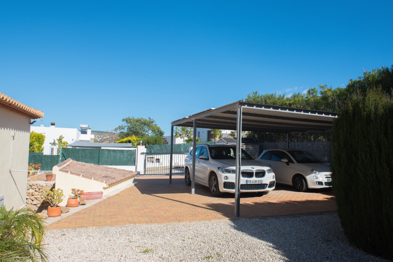 Espace de stationnement couvert avec abri métallique, sol en pavés de terre cuite et bordures en gravier blanc sous un ciel dégagé.