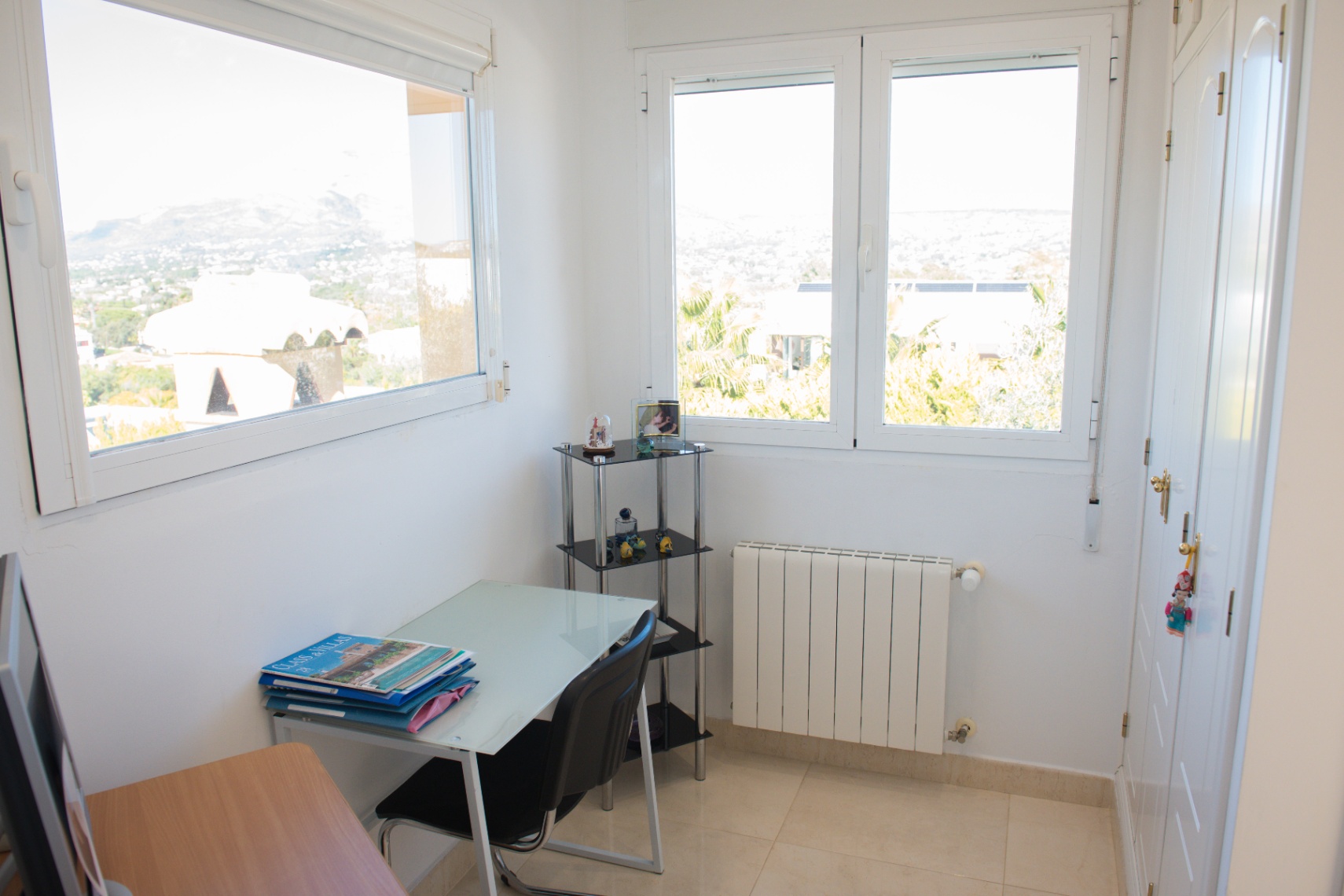 Espace de travail lumineux avec bureau en verre, placards intégrés blancs et double fenêtres offrant une vue panoramique sur les montagnes et les environs.