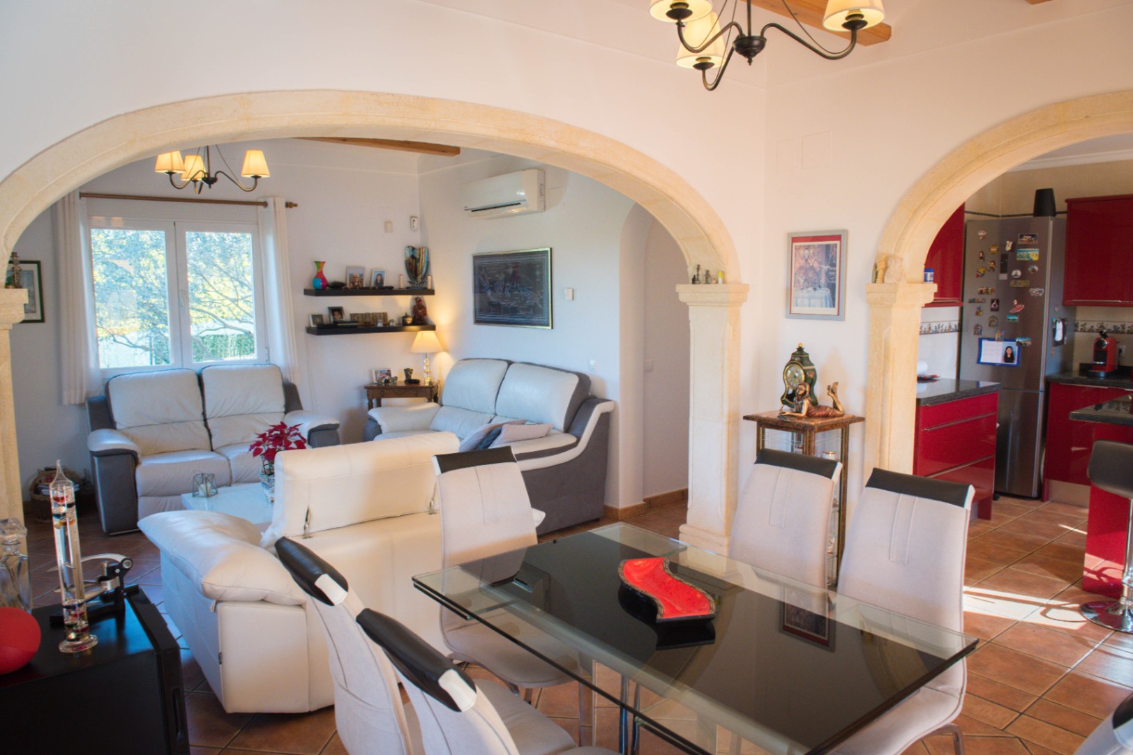 Espace de vie ouvert avec arches en pierre, salle à manger avec table en verre, canapés en cuir et cuisine moderne aux meubles rouges et sol en terre cuite.