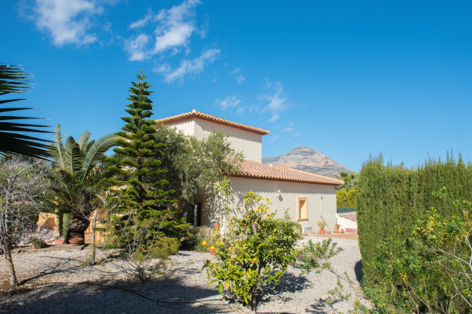 Extérieur de villa méditerranéenne avec toit en terre cuite et jardin de gravier composé d'agrumes, de palmiers et de cyprès sur fond de montagne.