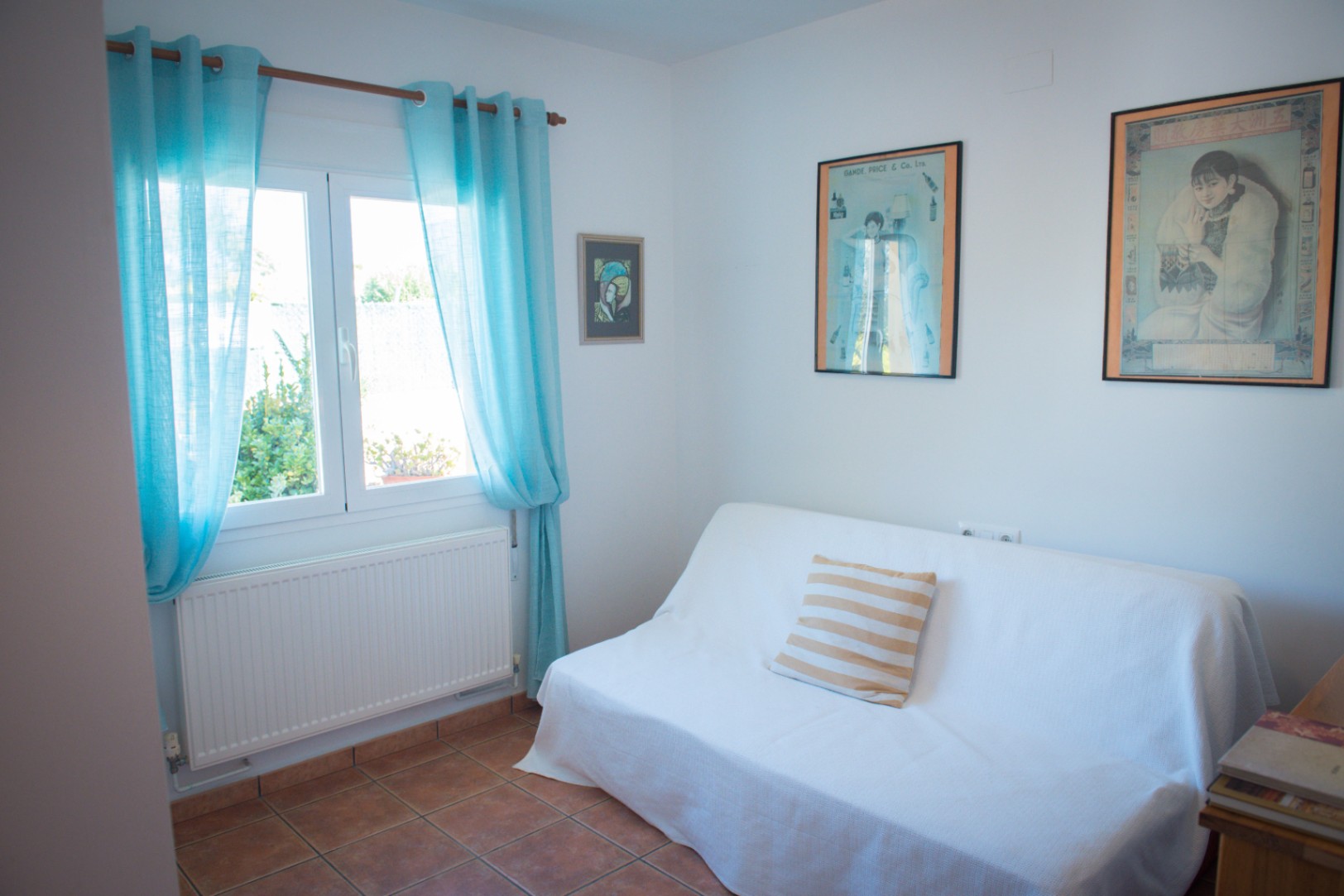 Pièce lumineuse avec sol en terre cuite, canapé blanc orné d'un coussin rayé et fenêtre avec rideaux turquoise apportant une lumière naturelle.