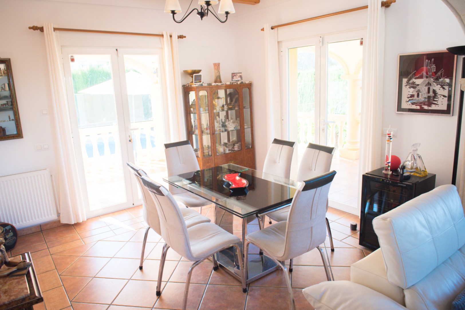 Salle à manger avec table en verre et chaises blanches. Le sol en terre cuite et les doubles portes vitrées offrent une luminosité naturelle optimale.