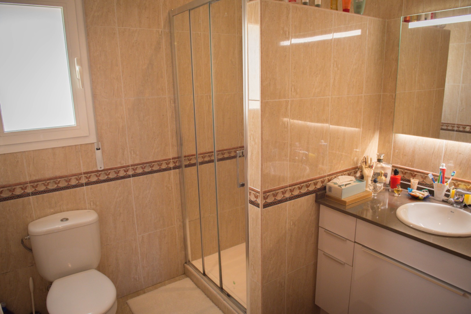 Salle de bain aux murs carrelés beige avec frise, comprenant une douche vitrée, des toilettes blanches et un meuble vasque éclairé.