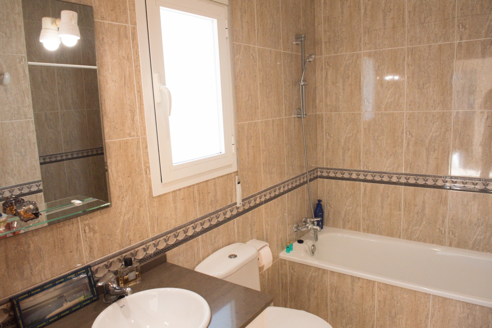 Salle de bain carrelée en beige avec baignoire, lavabo intégré, miroir avec éclairage et fenêtre blanche assurant une ventilation naturelle.