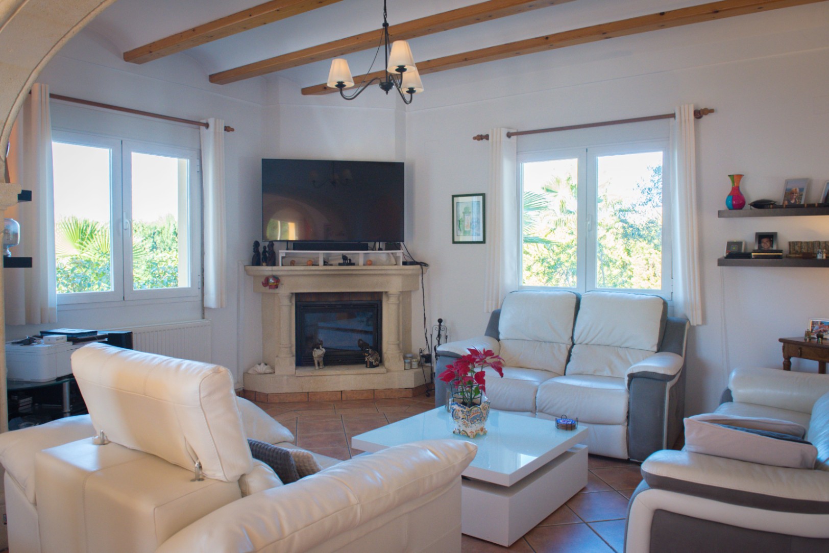 Salon avec poutres apparentes en bois, cheminée d'angle en pierre surmontée d'un téléviseur, canapés en cuir blanc et sol en terre cuite.