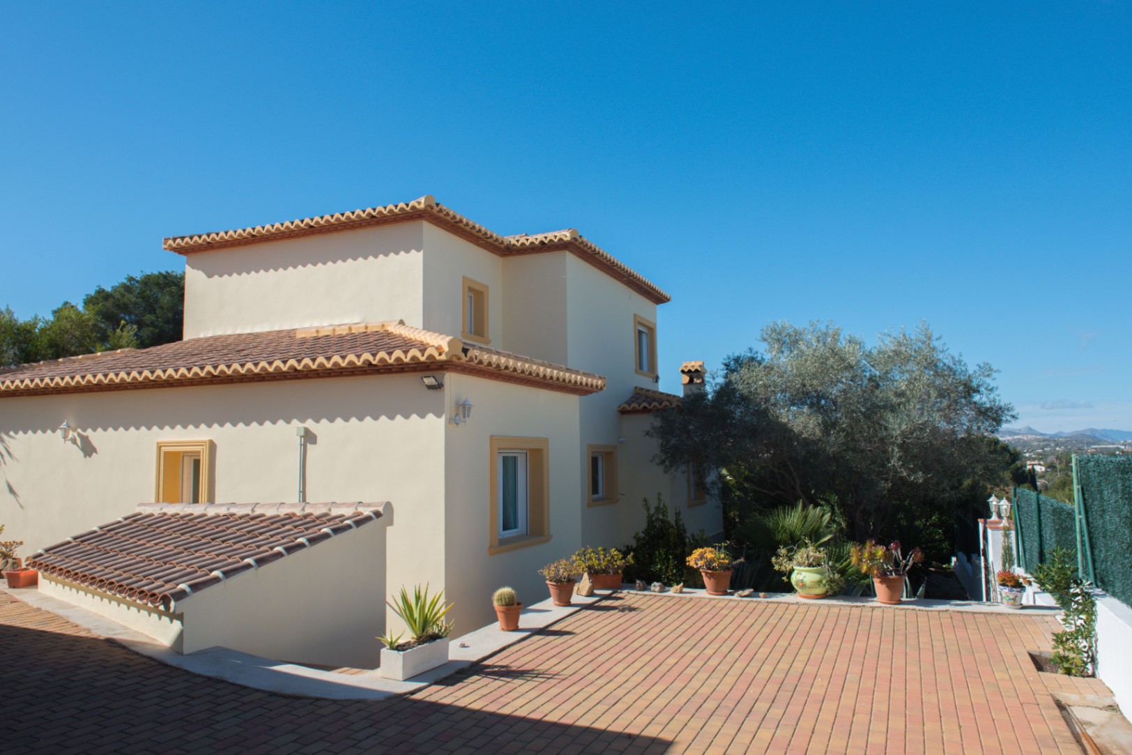 Vue extérieure d'une villa méditerranéenne aux murs crème et toits en tuiles, complétée par une terrasse pavée de briques et des plantes en pots.