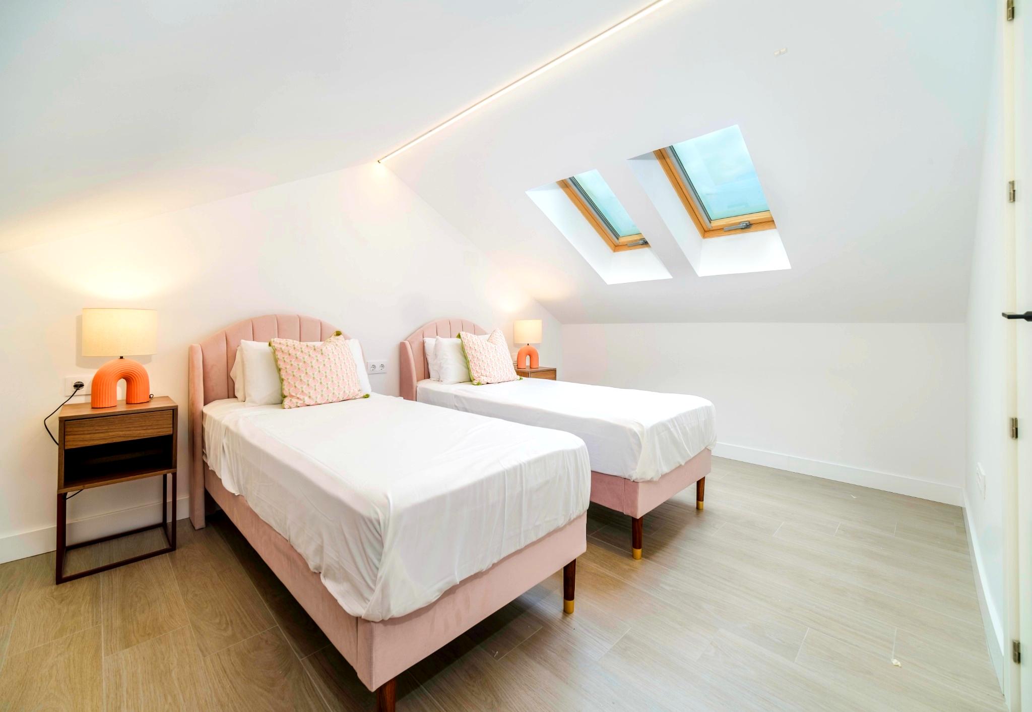 Chambre mansardée avec deux lits simples en velours rose, doubles fenêtres de toit et un éclairage LED intégré au plafond blanc épuré.