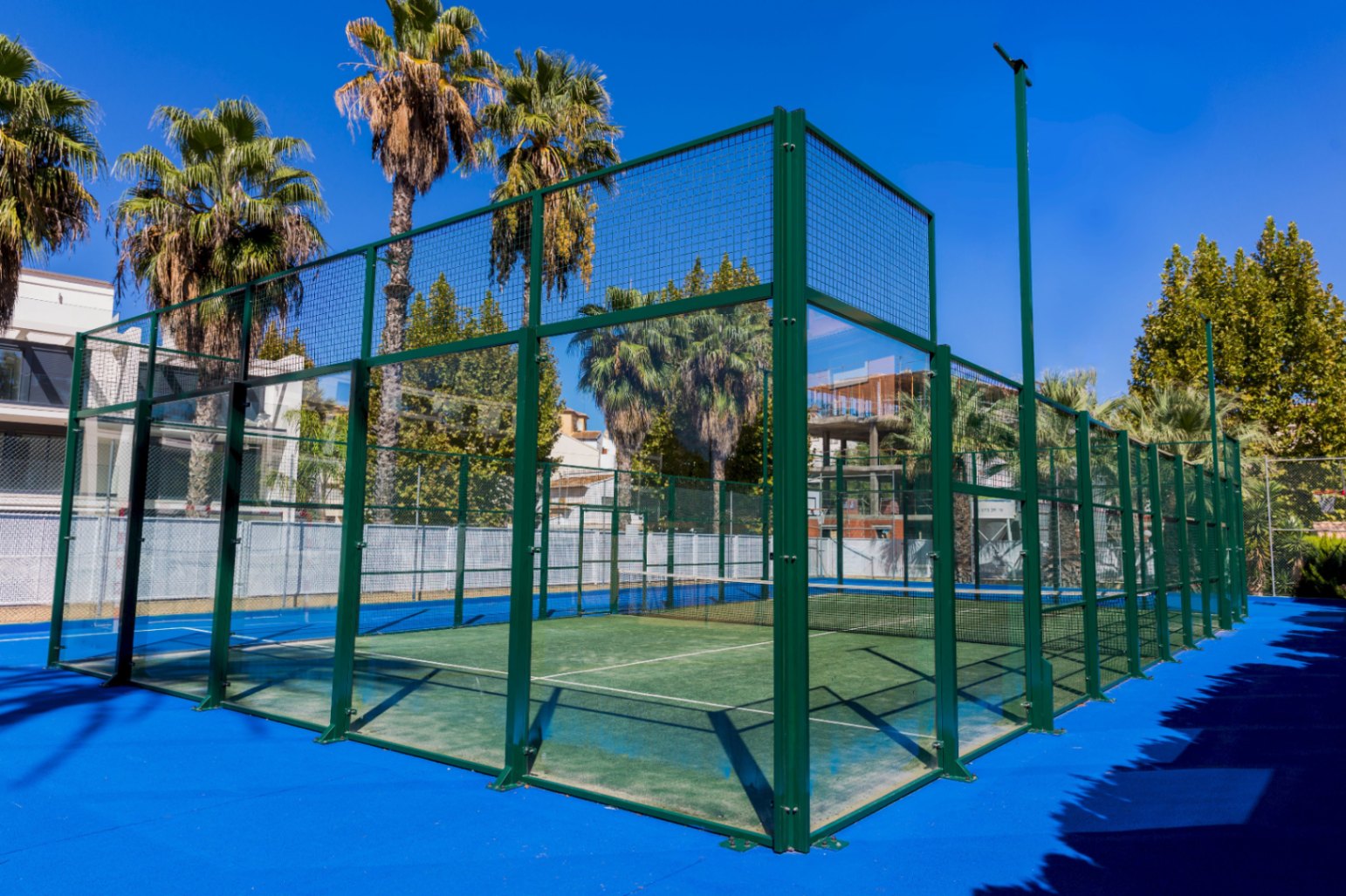 Court de padel extérieur avec gazon synthétique vert, parois en verre trempé et structure métallique, entouré de palmiers sous un ciel dégagé.