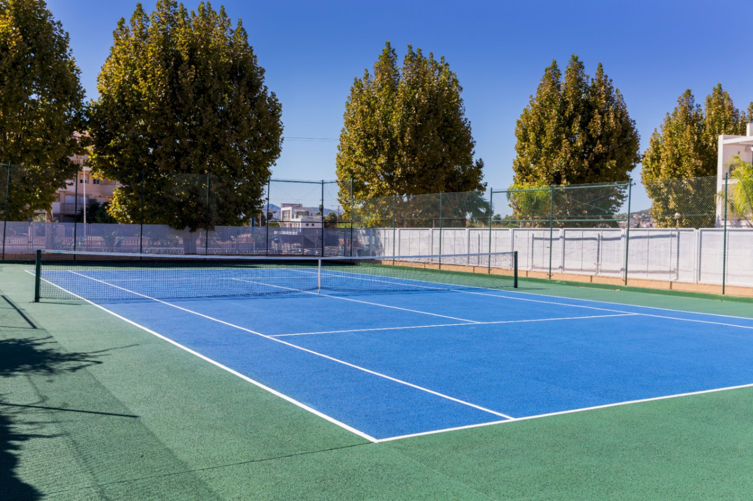 Court de tennis extérieur en surface dure avec zone de jeu bleue et bordure verte, entouré d'une clôture haute et de grands arbres sous un ciel dégagé.