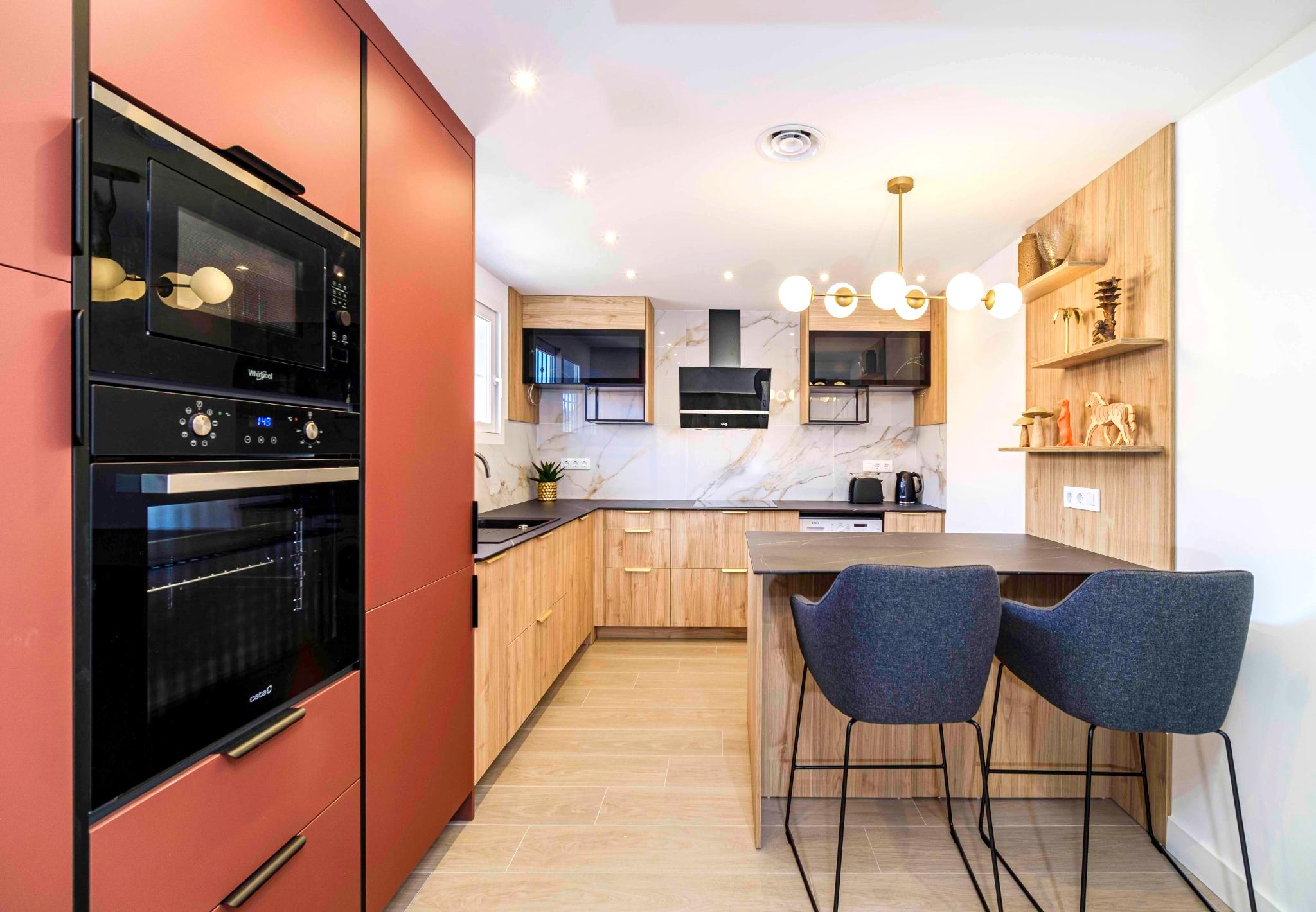 Cuisine moderne avec colonnes terracotta, meubles en bois clair, crédence effet marbre et un îlot dînatoire avec tabourets bleus et luminaire design doré.