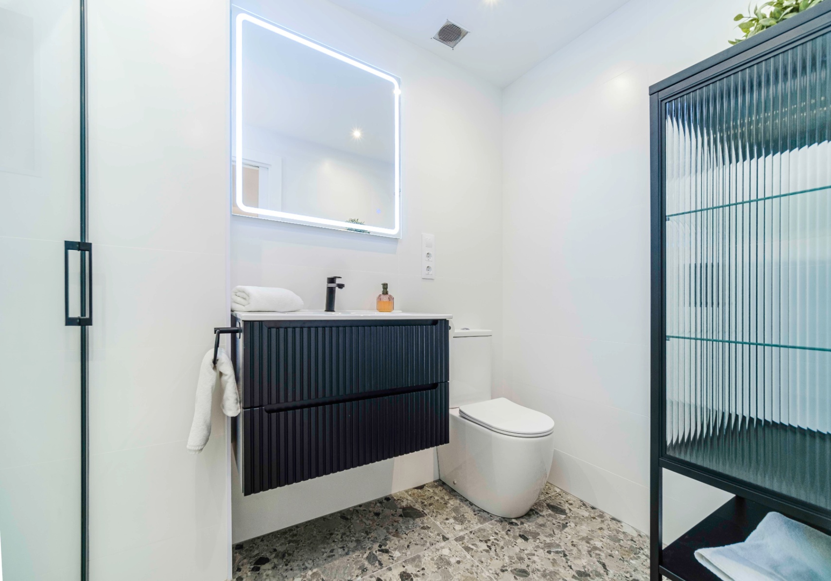 Salle de bain moderne avec meuble vasque suspendu noir cannelé, miroir à LED et sol en terrazzo gris élégant.
