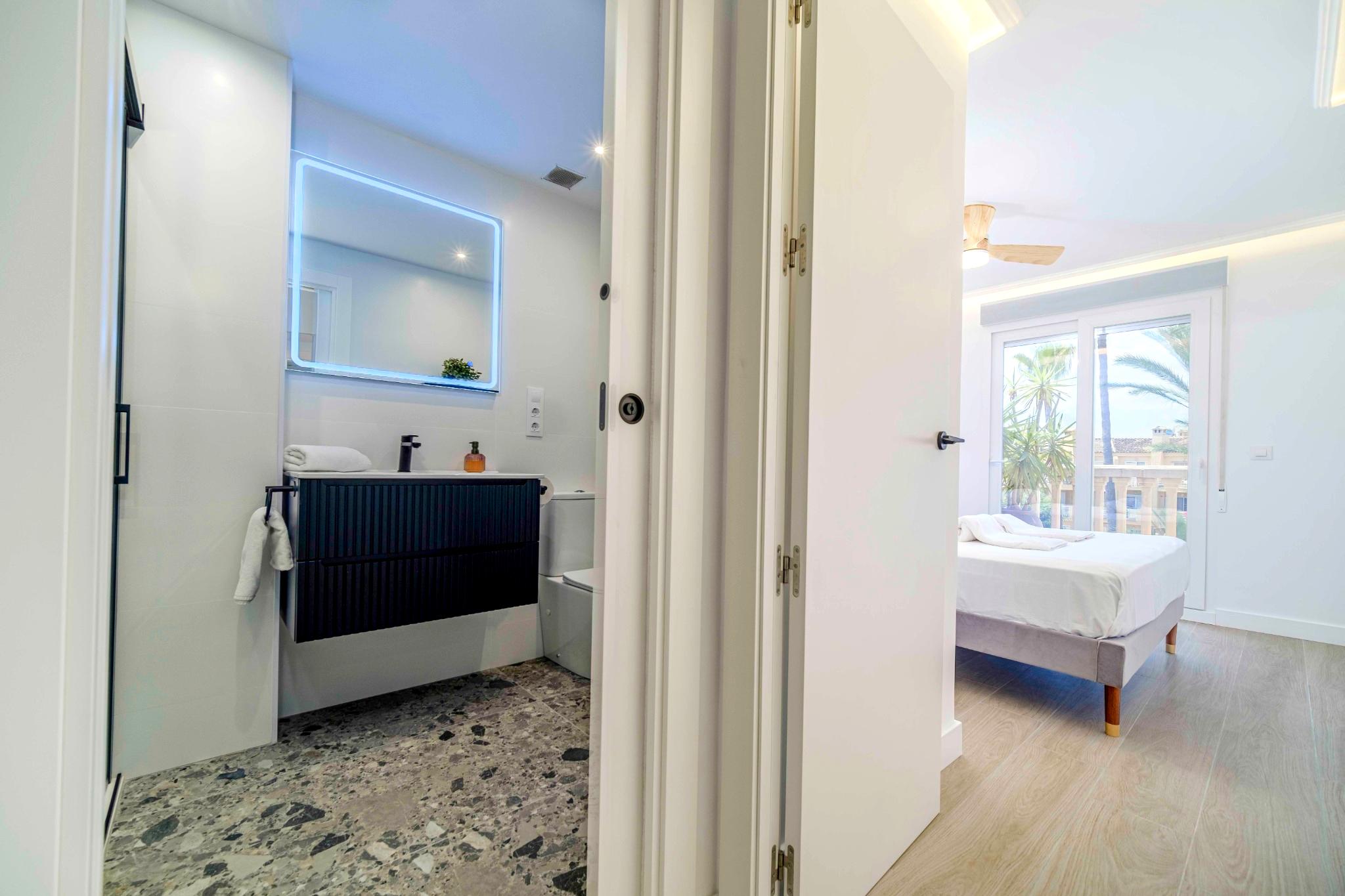 Salle de bain moderne avec sol en terrazzo et meuble noir, attenante à une chambre lumineuse offrant un accès au balcon et des vues extérieures.