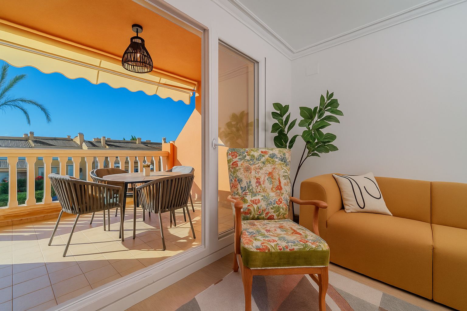 Salon avec canapé moutarde et fauteuil à fleurs s'ouvrant sur une terrasse privée avec coin repas extérieur, store banne jaune et vue dégagée.