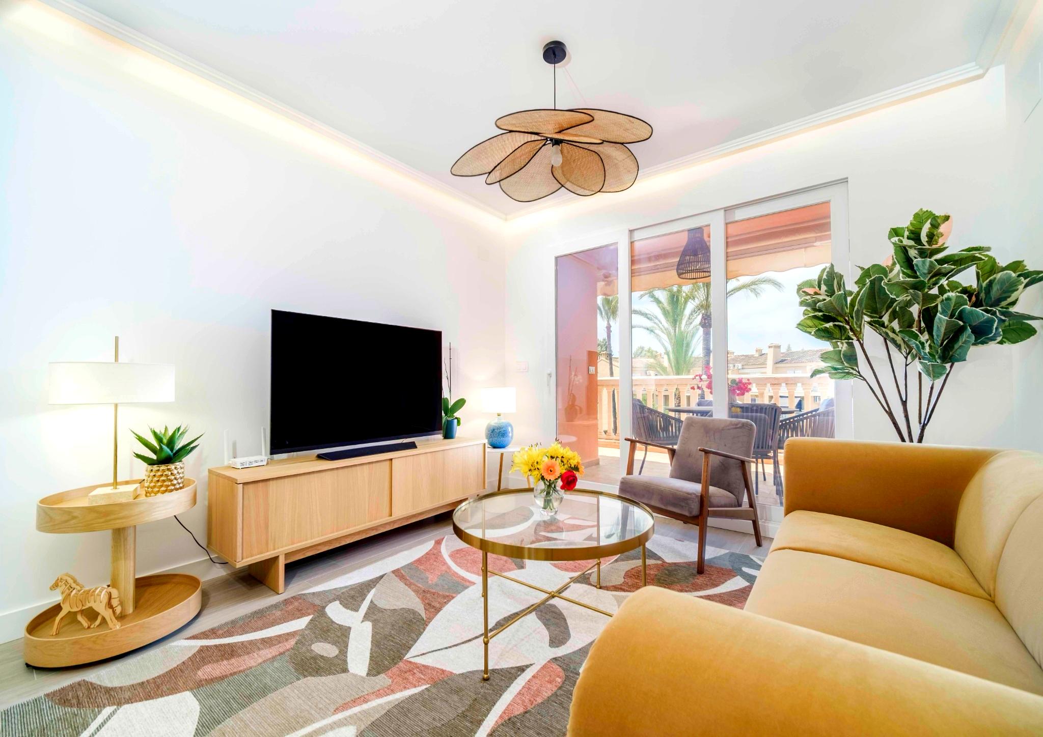 Salon contemporain avec canapé jaune, luminaire en forme de pétales et grandes baies vitrées ouvrant sur une terrasse bordée de palmiers.