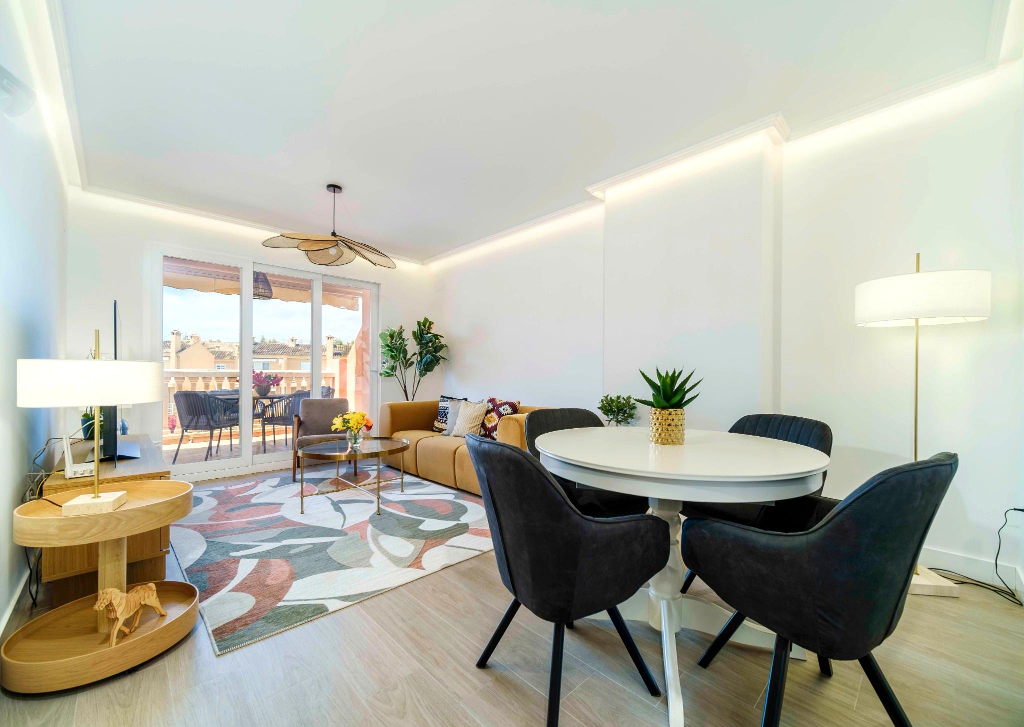 Séjour ouvert avec canapé moutarde, table à manger ronde et éclairage LED intégré. De grandes baies vitrées s'ouvrent sur une terrasse meublée.