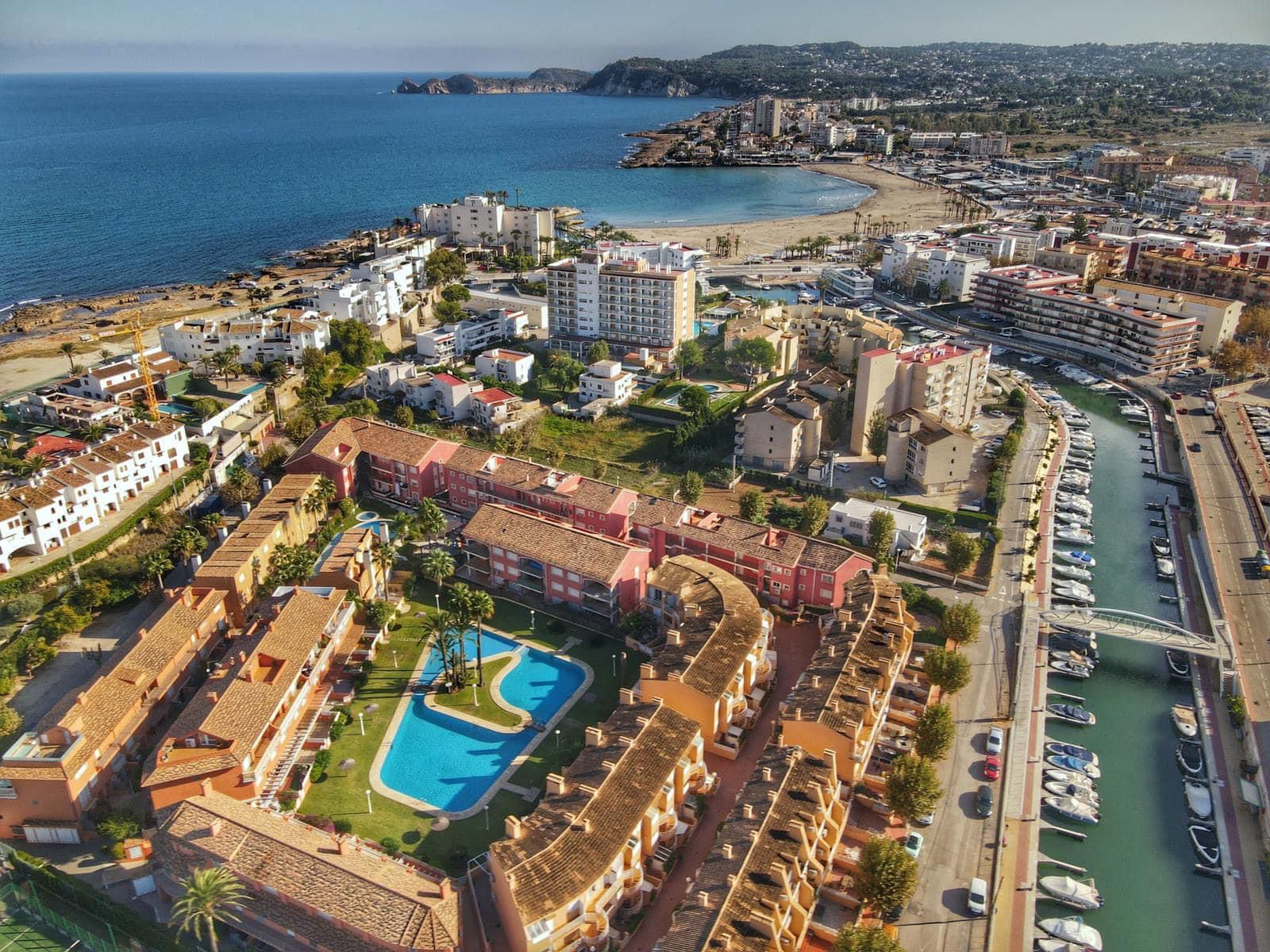 Vue aérienne d'une résidence avec piscine paysagère et toits en terre cuite, située entre un canal de plaisance et la plage de sable fin.