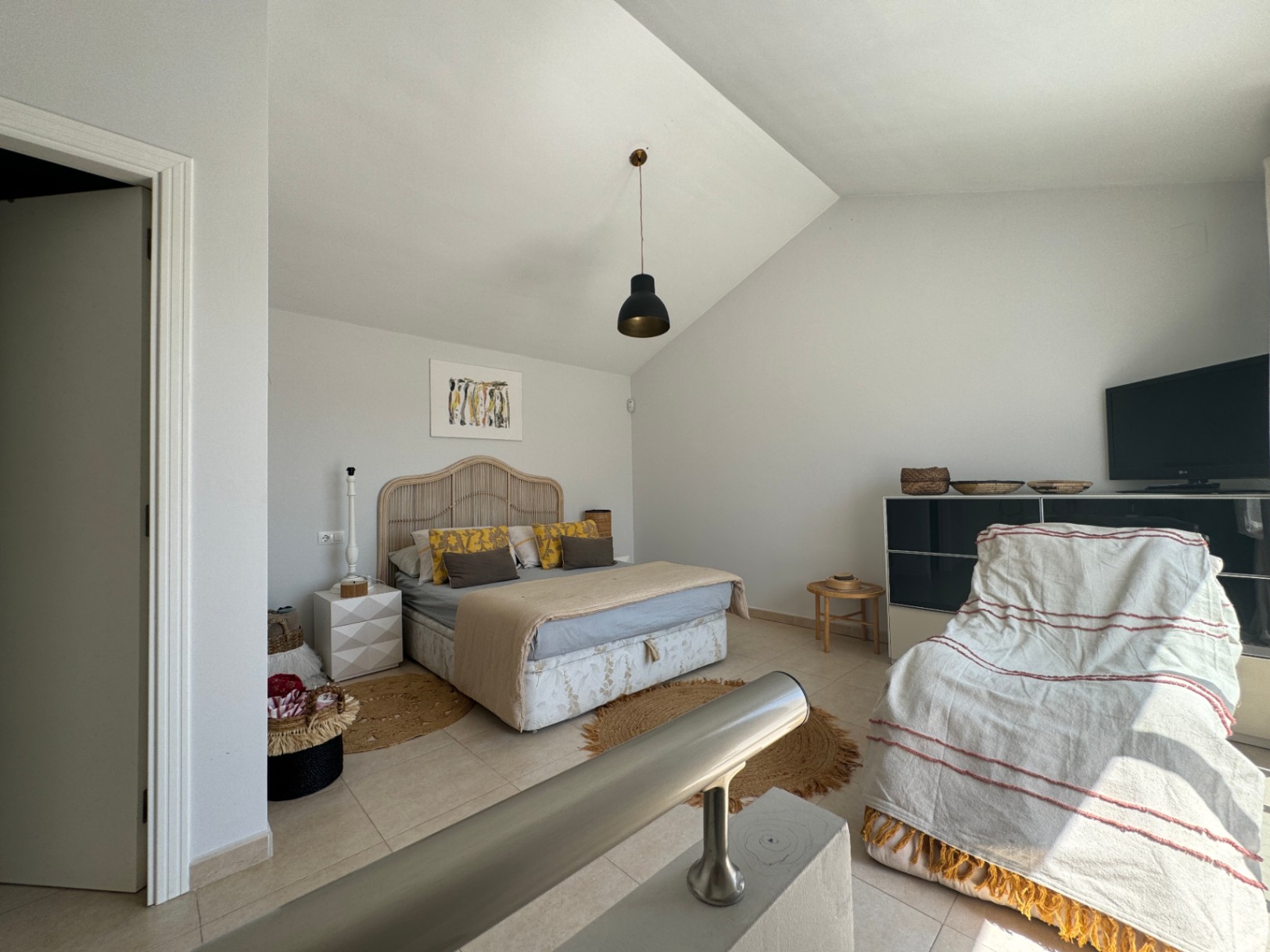 Chambre avec plafond mansardé, tête de lit en rotin et suspension noire. Elle dispose d'un sol en carrelage, de tapis en jute et d'une rampe métallique au premier plan.