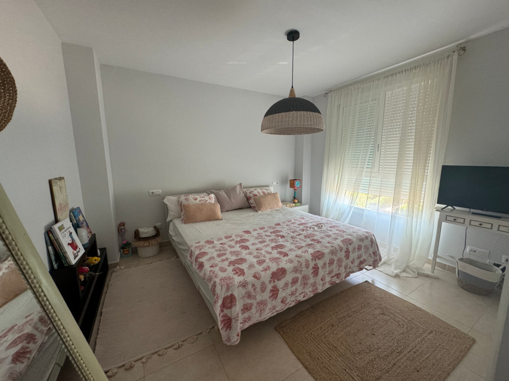 Chambre lumineuse avec lit double, parure de lit aux motifs corail, suspension en fibres tressées et grande fenêtre avec voilages blancs.