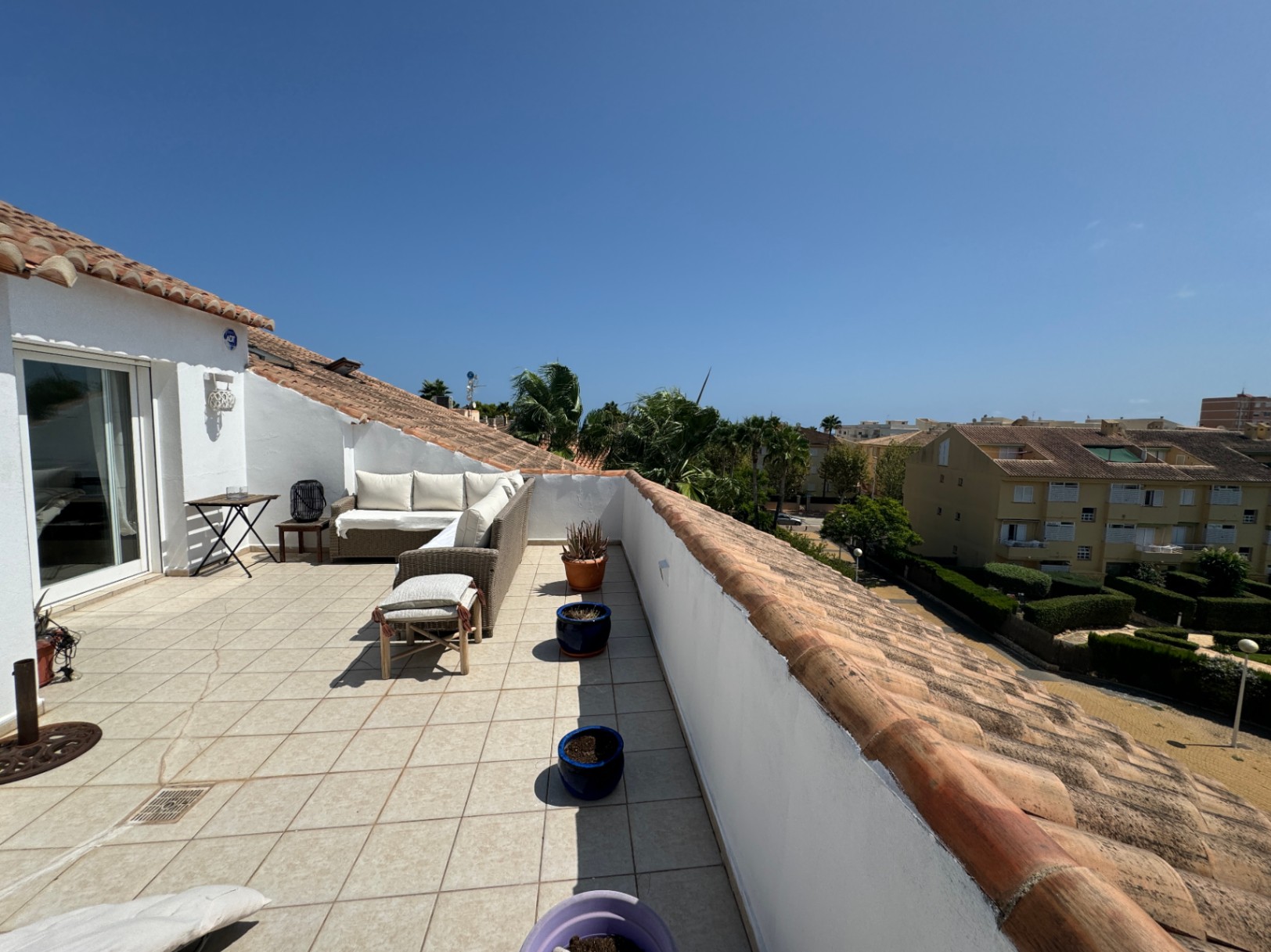 Grande terrasse de toit avec carrelage clair, canapé d'angle en rotin et coussins blancs, offrant une vue sur les environs résidentiels et la végétation.