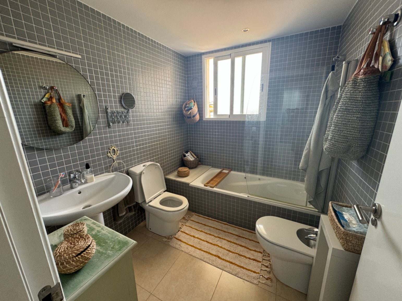 Salle de bain avec carrelage en mosaïque grise, comprenant un lavabo sur colonne, un miroir circulaire et une baignoire avec paroi vitrée éclairée par une fenêtre.