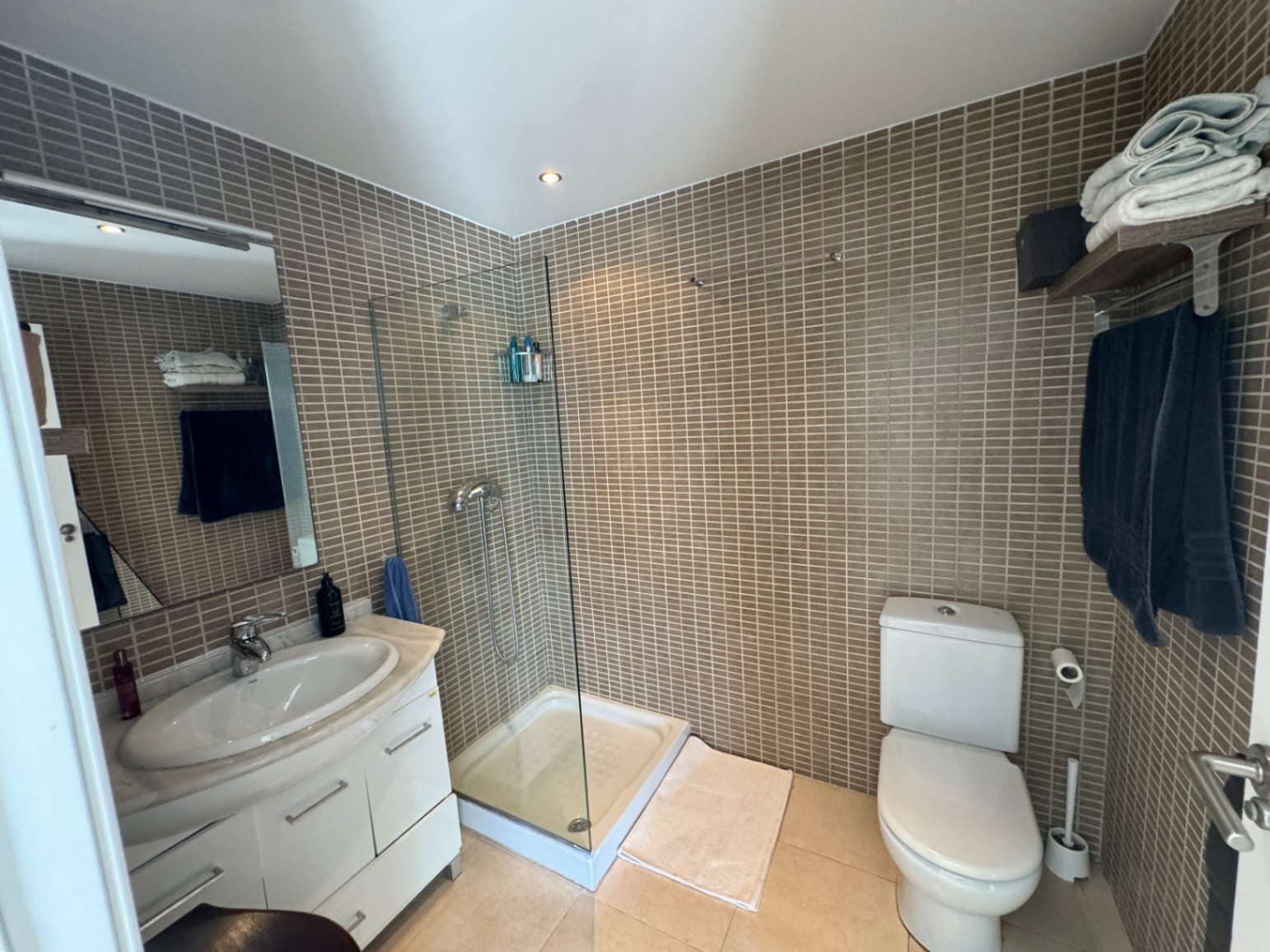 Salle de bain avec douche vitrée, murs en mosaïque brune et meuble vasque blanc avec plan de travail en marbre et éclairage encastré.