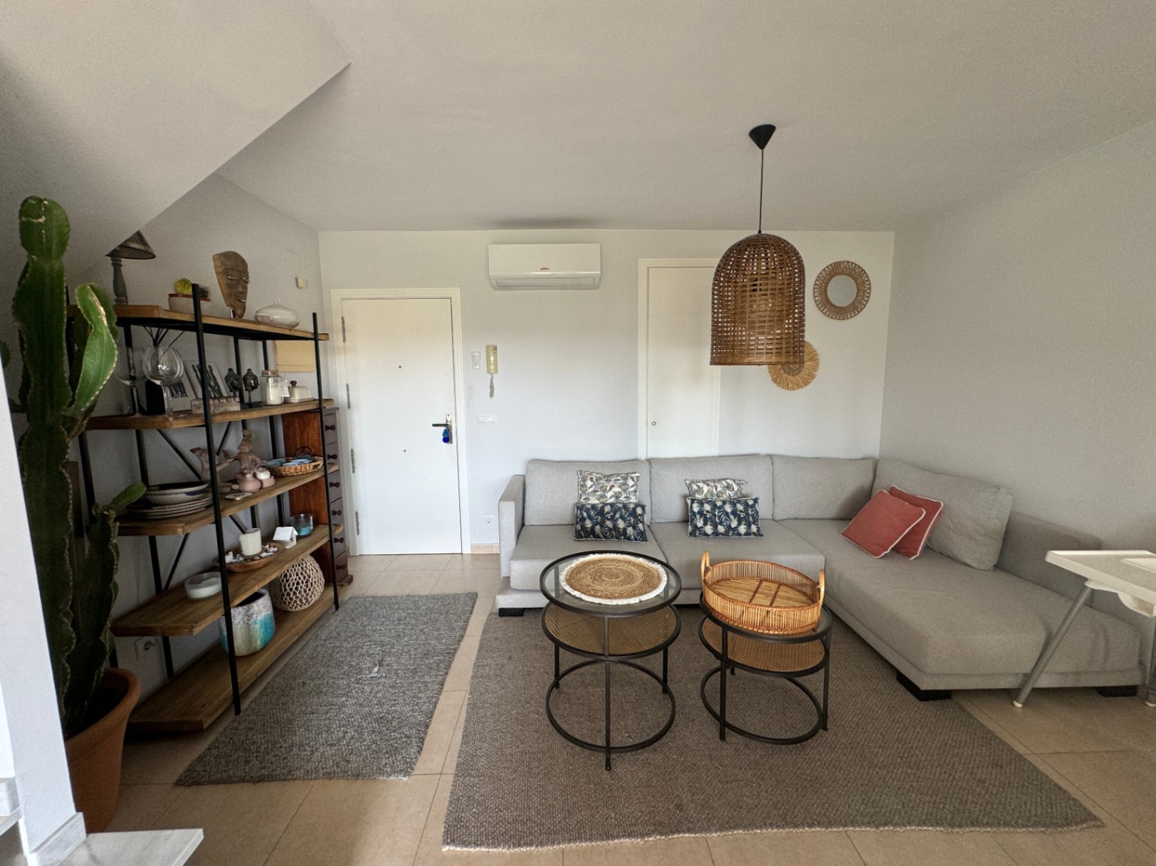 Salon avec canapé d'angle gris, suspension en rotin et étagère industrielle en bois. Les tons neutres et le sol carrelé apportent une atmosphère sereine et moderne.