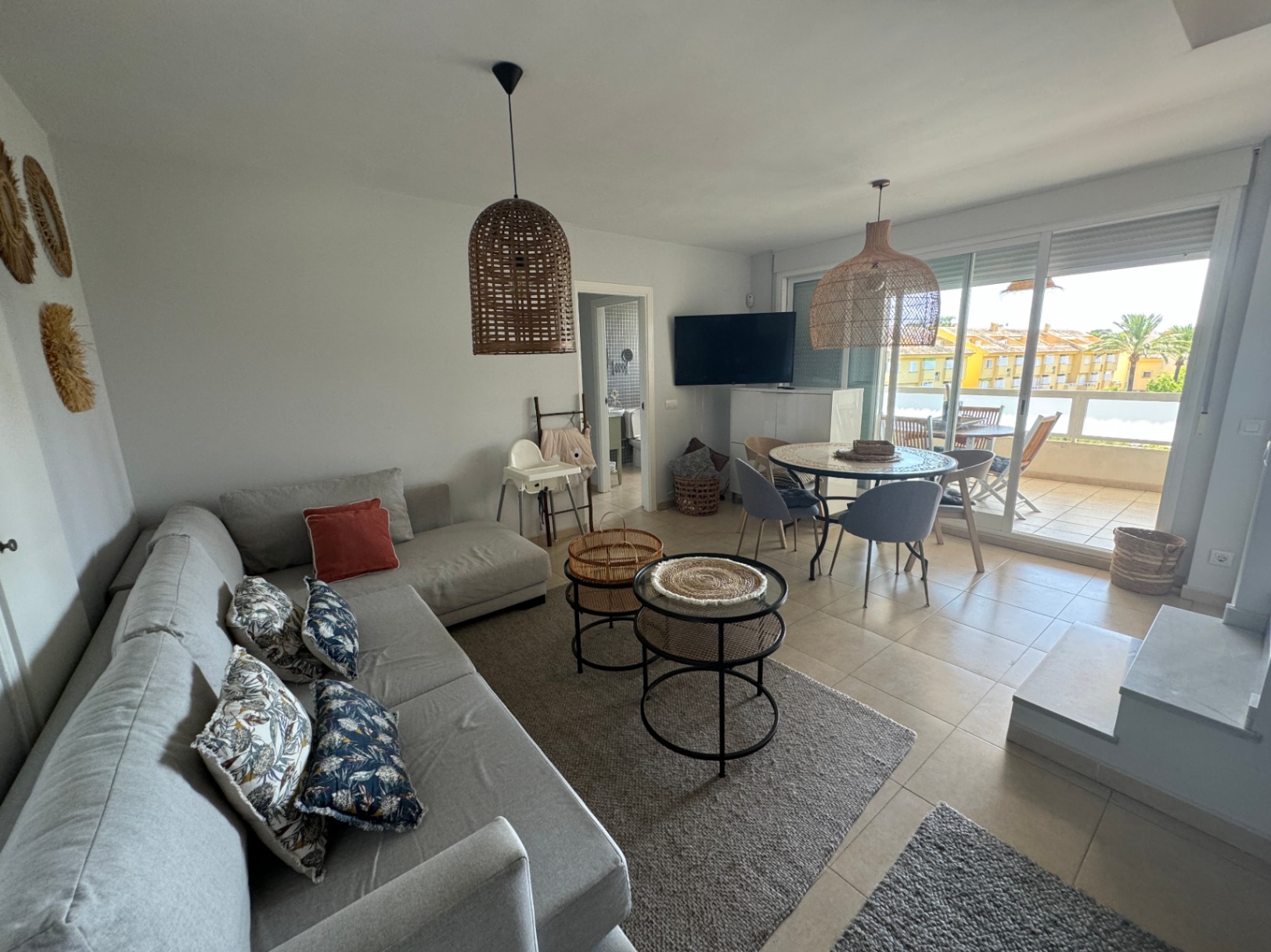 Salon-salle à manger lumineux avec canapé d'angle gris, suspensions en rotin et table ronde. Les baies vitrées s'ouvrent sur une terrasse privée avec vue dégagée.