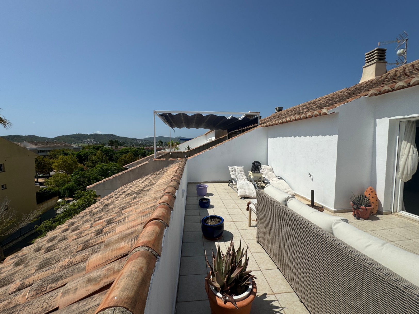 Terrasse de toit allongée avec canapé en osier gris, chaises longues blanches et carrelage en terre cuite, offrant une vue sur les collines environnantes.