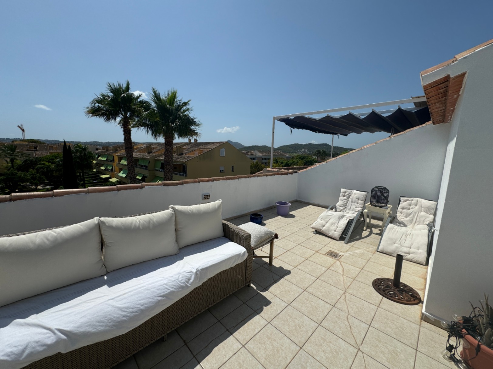 Terrasse sur le toit avec sol en carrelage, canapé en osier, chaises longues et store rétractable. Vue sur les palmiers et le paysage environnant.