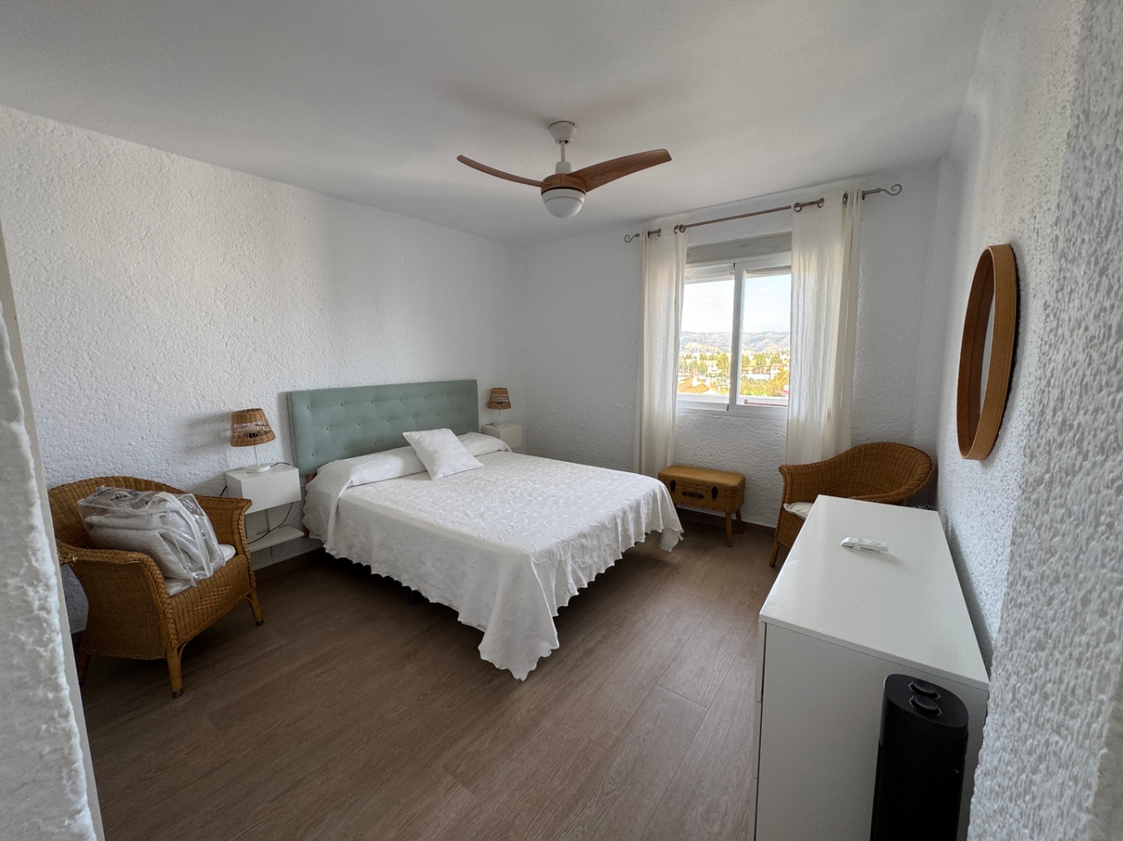 Chambre aux murs texturés blancs avec lit double, tête de lit vert menthe, fauteuils en osier et ventilateur de plafond à pales en bois.
