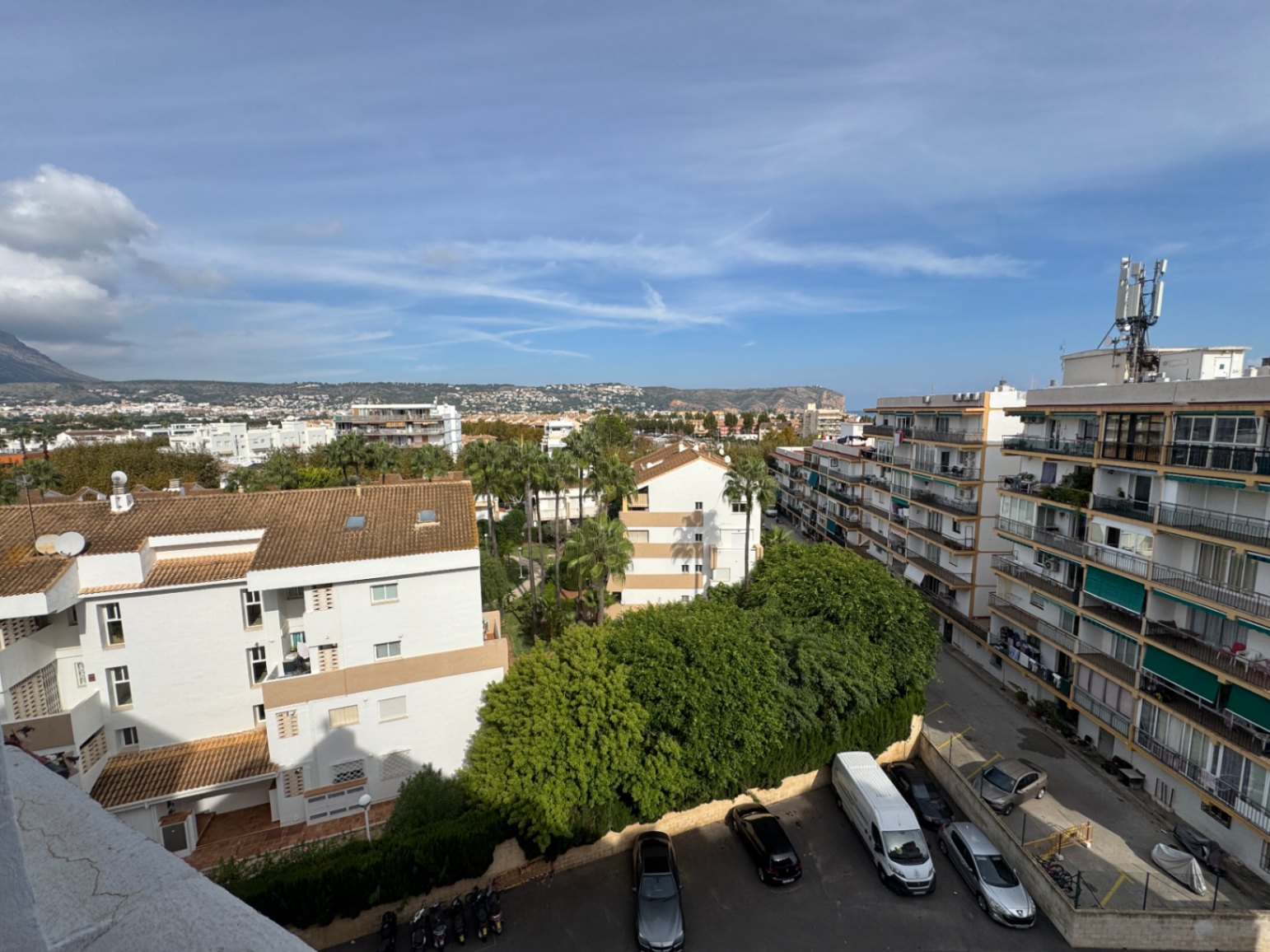 Vue surélevée présentant des bâtiments résidentiels, des palmiers matures et un parking, avec des collines côtières à l'horizon.