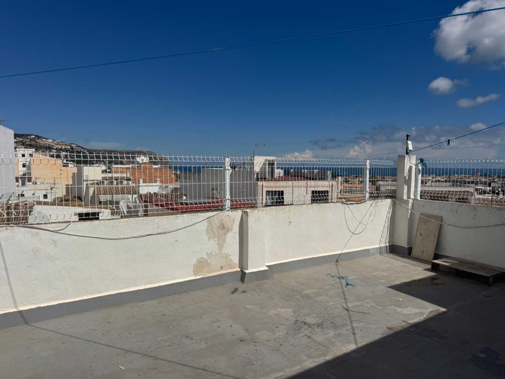 Terrasse sur le toit avec sol carrelé et murs blancs surmontés d'un grillage, offrant une vue sur la ville, les collines environnantes et la mer.