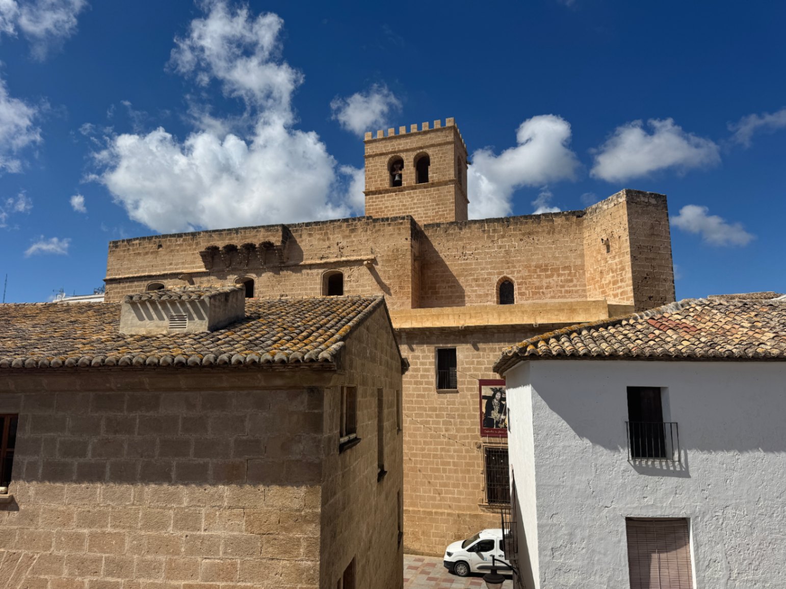 Vue d'une église fortifiée en pierre avec un clocher crénelé, entourée de maisons méditerranéennes traditionnelles aux toits de tuiles sous un ciel bleu.