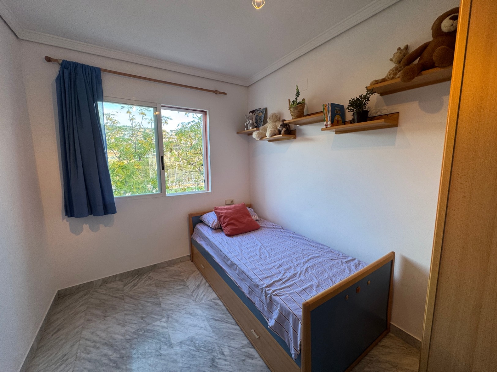Chambre simple avec sol en marbre, étagères murales en bois et fenêtre coulissante vue sur jardin. Le lit dispose de tiroirs de rangement intégrés.