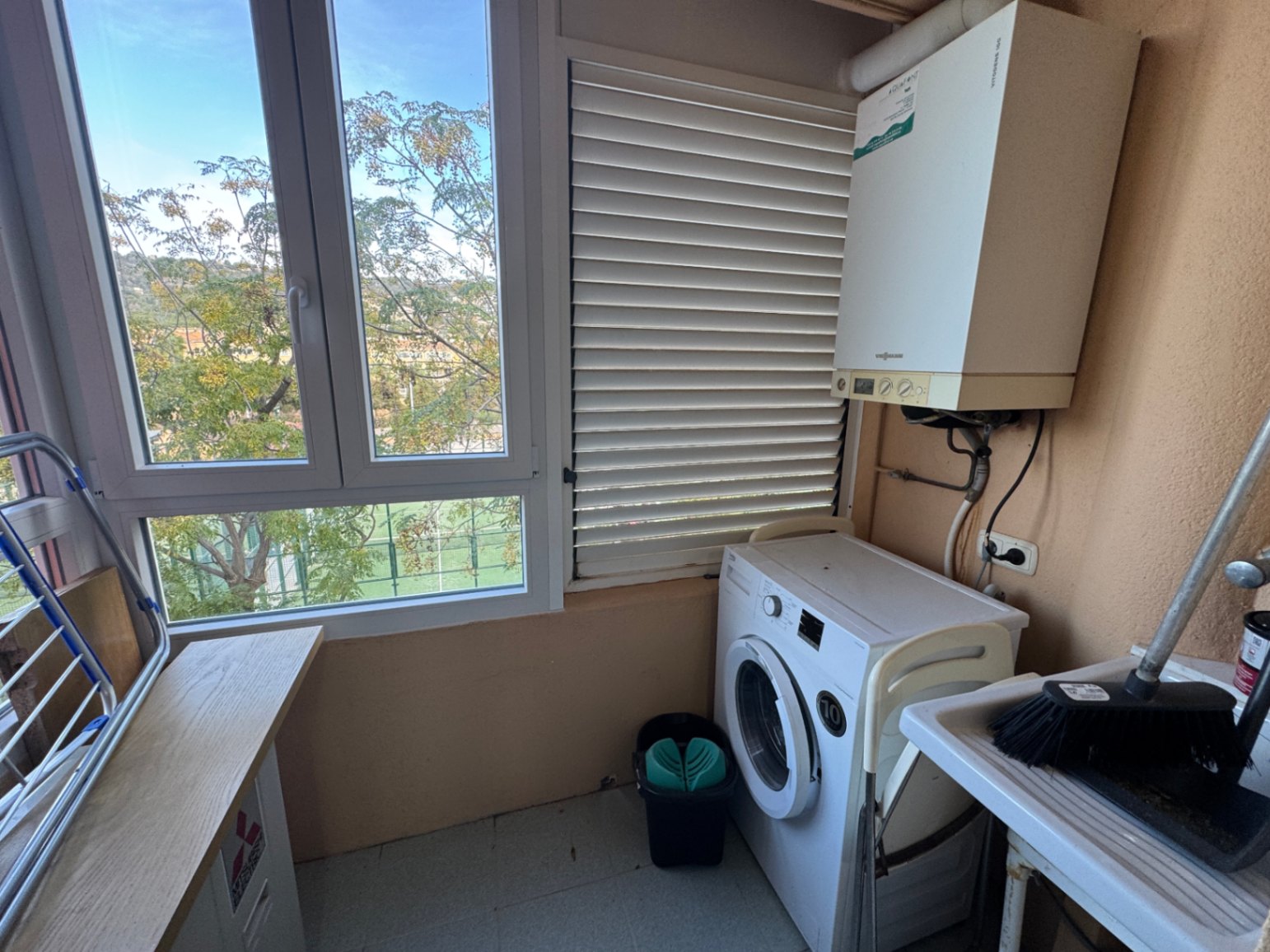 Espace buanderie fonctionnel avec lave-linge, chaudière murale et évier. Les grandes fenêtres offrent une luminosité naturelle et une vue dégagée sur l'extérieur.