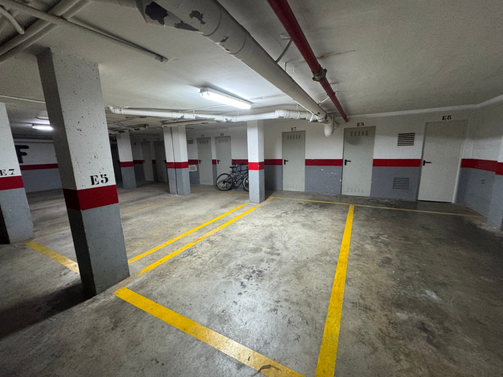 Parking souterrain avec sol en béton, marquages jaunes et portes de box numérotées. Comprend des tuyauteries apparentes et un éclairage fonctionnel au plafond.