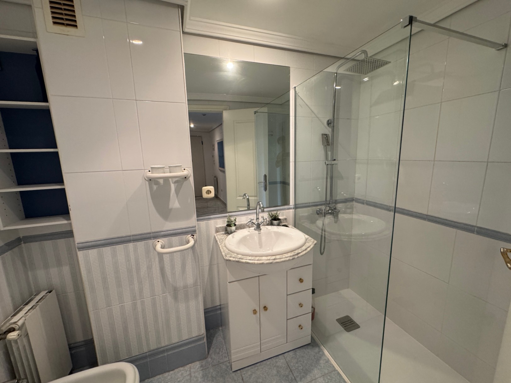 Salle de bain avec douche à l'italienne vitrée, meuble vasque blanc avec plan en marbre et étagères encastrées. Les murs sont parés de carreaux blancs et rayés.