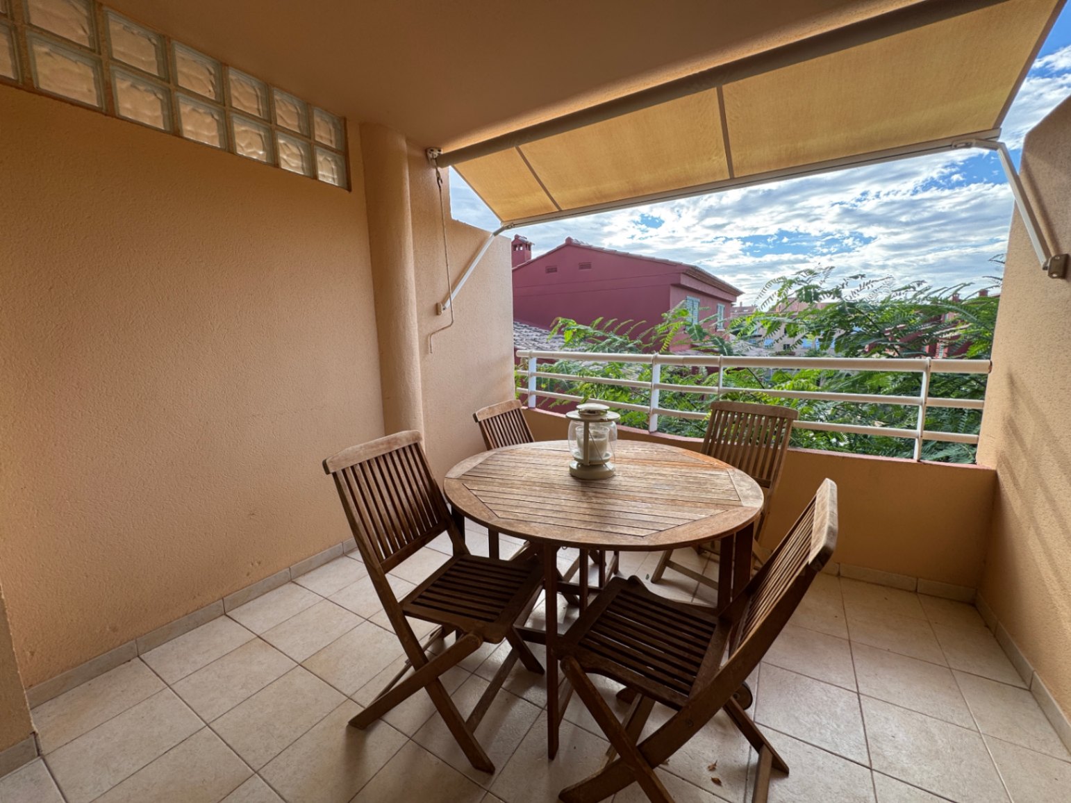 Terrasse couverte avec ensemble repas en bois, sol carrelé et store banne offrant une vue sur la végétation et les bâtiments environnants.