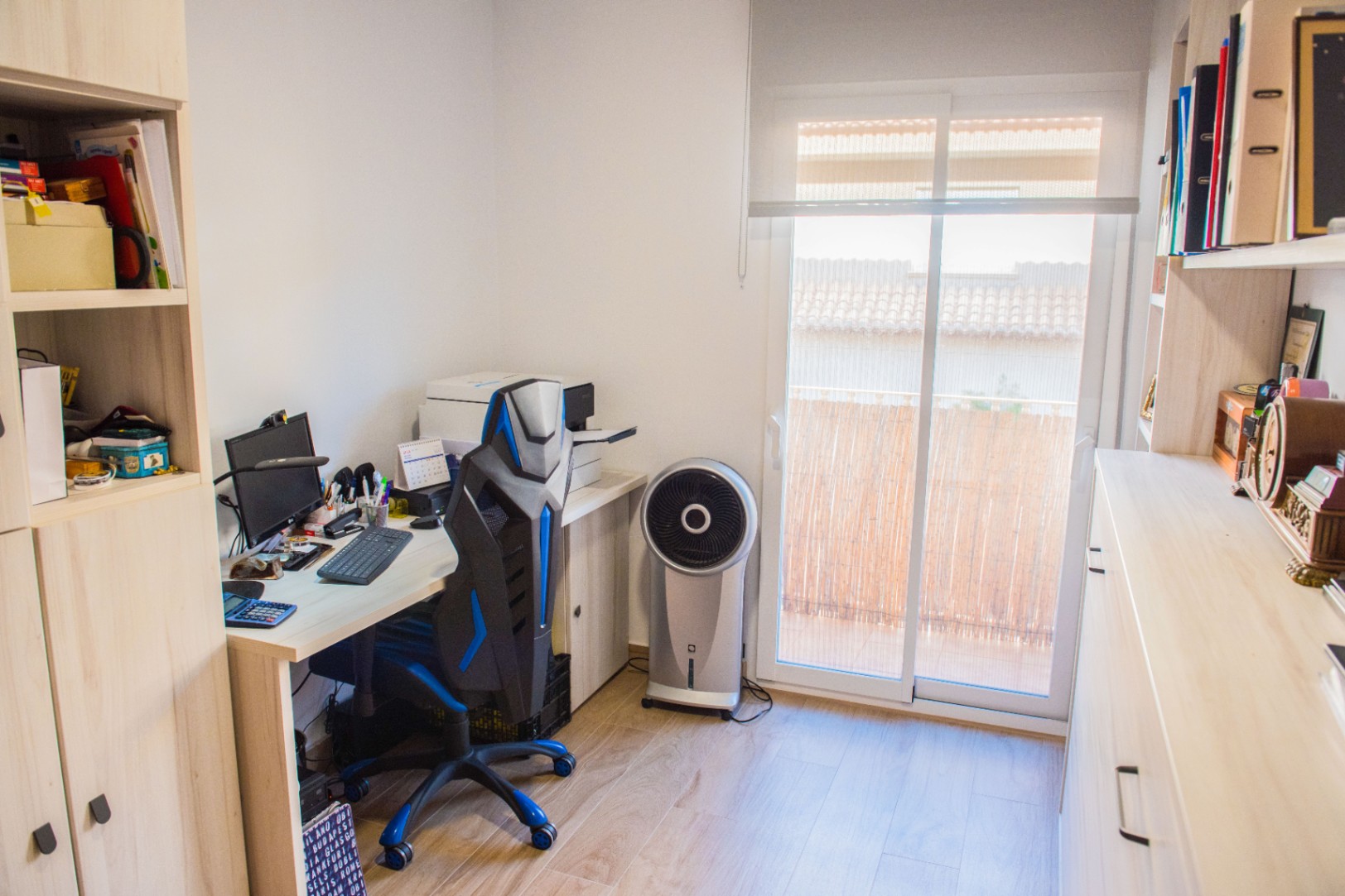 Bureau avec mobilier en bois clair, chaise ergonomique et étagères intégrées. Une grande porte coulissante en verre donne accès à un balcon baigné de lumière naturelle.
