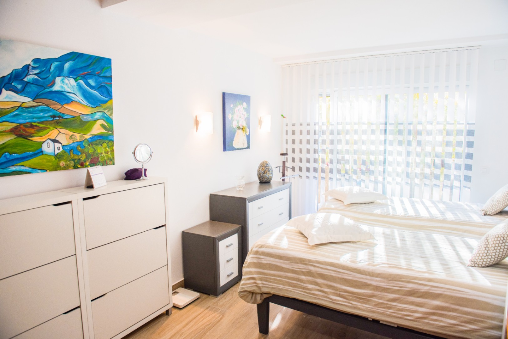 Chambre lumineuse avec deux lits simples, commodes minimalistes et tableaux colorés. La lumière naturelle filtre à travers des stores verticaux sur un sol imitation bois.