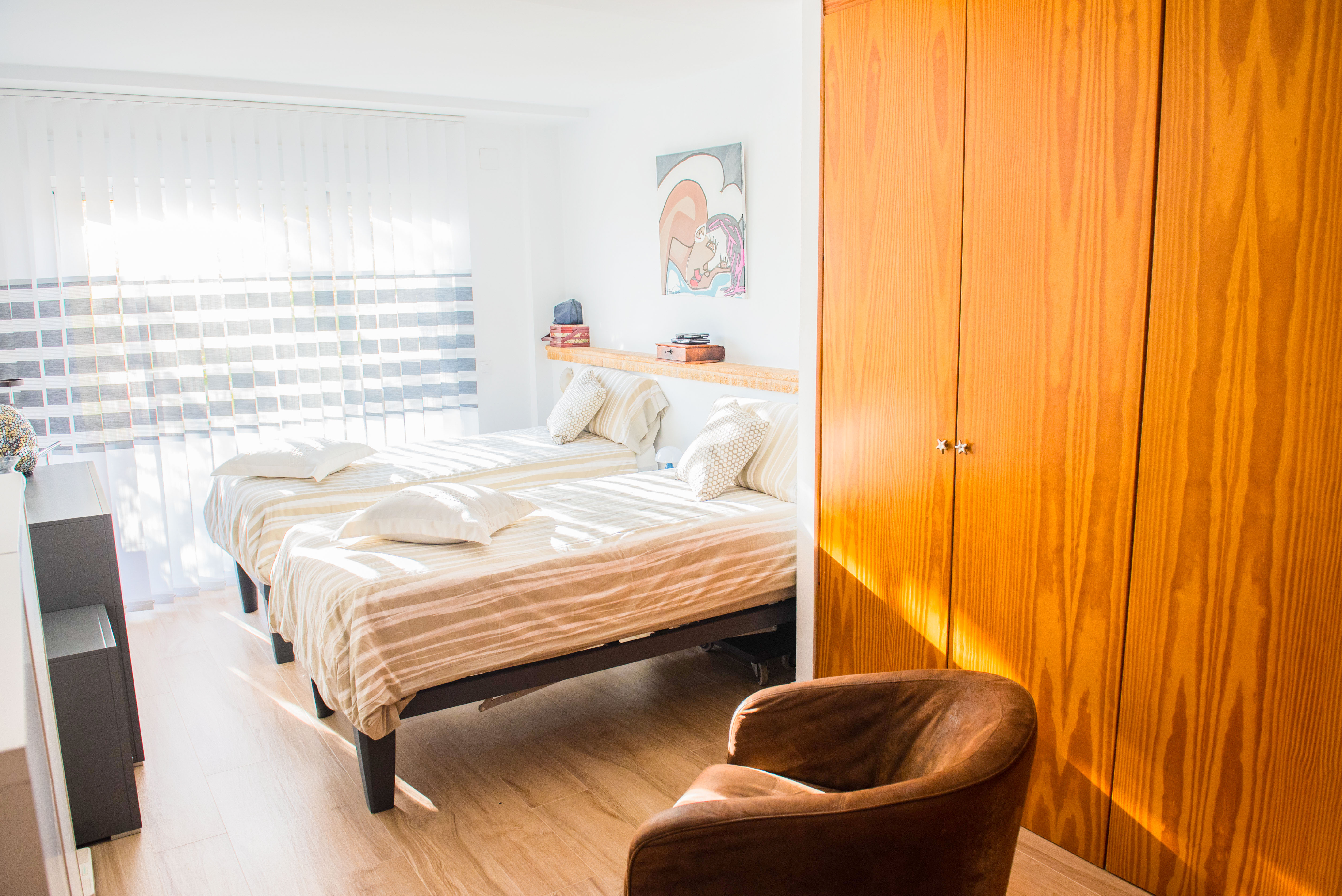 Chambre lumineuse avec deux lits simples, une armoire en bois naturel et un sol clair. La lumière naturelle pénètre par les stores verticaux, éclairant la pièce. Chambre lumineuse avec deux lits simples, une armoire en bois naturel et un sol clair. La lumière naturelle pénètre par les stores verticaux, éclairant la pièce.