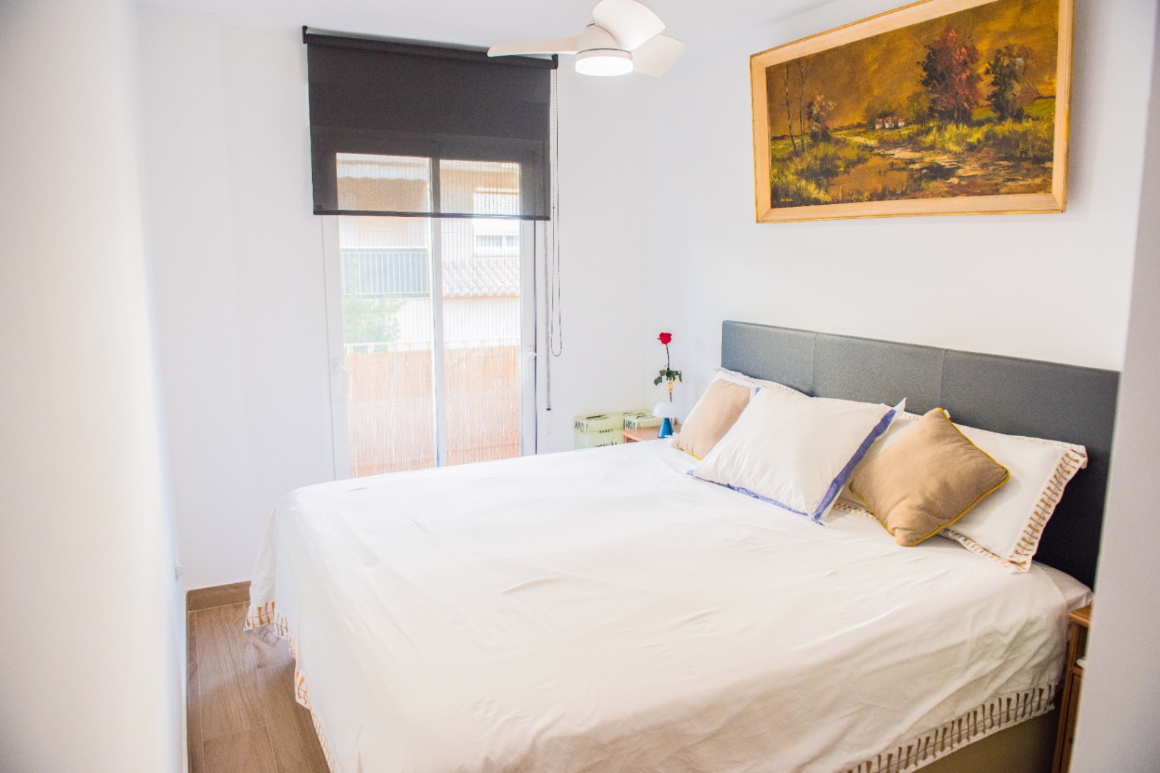 Chambre lumineuse avec tête de lit grise, ventilateur de plafond et store noir. Un tableau paysager à l'huile décore le mur au-dessus du lit.