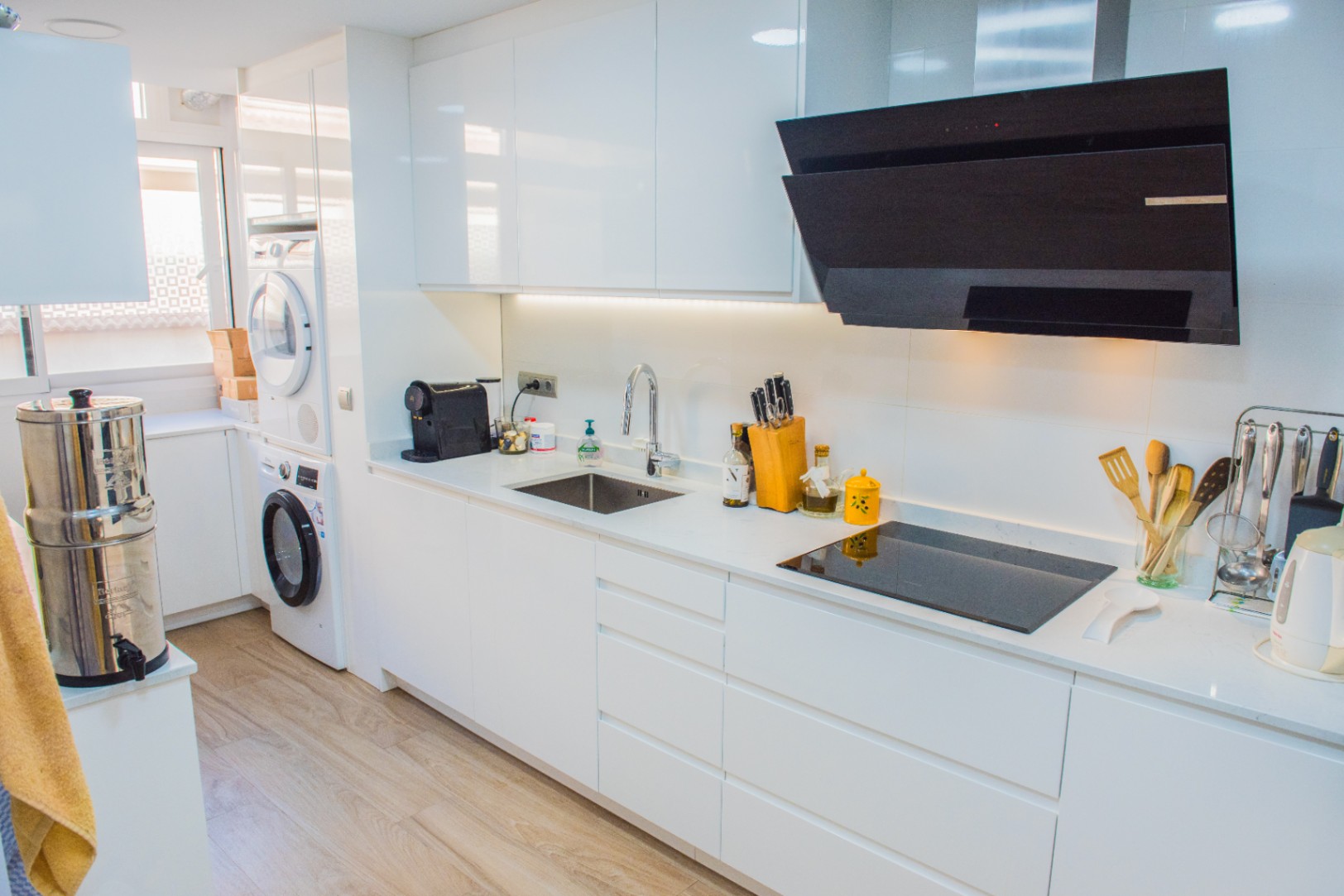 Cuisine blanche minimaliste avec meubles brillants, plaque à induction, hotte noire et espace buanderie intégré avec lave-linge et sèche-linge.