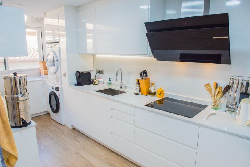Cuisine moderne avec armoires blanches laquées, plaque à induction noire et hotte inclinée. L'espace intègre un éclairage sous les meubles et des appareils de buanderie près de la fenêtre. Cuisine moderne avec armoires blanches laquées, plaque à induction noire et hotte inclinée. L'espace intègre un éclairage sous les meubles et des appareils de buanderie près de la fenêtre.
