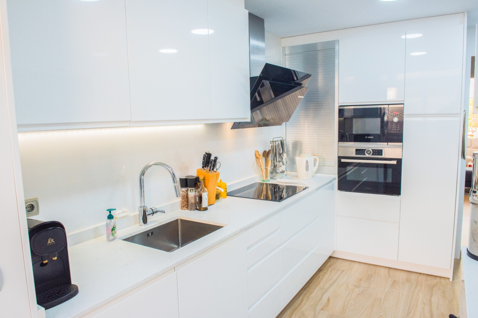 Cuisine moderne avec placards blancs brillants, électroménager Bosch encastré et plan de travail en quartz blanc équipé d'une plaque à induction et d'une hotte inclinée.