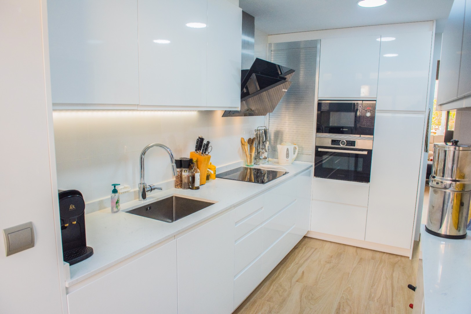 Cuisine moderne avec placards blancs brillants, électroménagers Bosch encastrés et plaque à induction. Comprend un plan de travail en quartz blanc et un sol en bois clair.