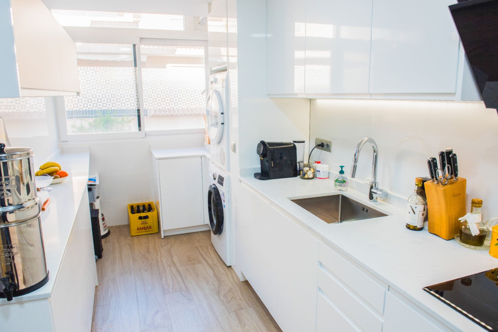 Cuisine moderne en longueur avec placards blancs sans poignées, espace buanderie intégré et plan de travail blanc. Éclairage LED sous les meubles et sol en bois clair.