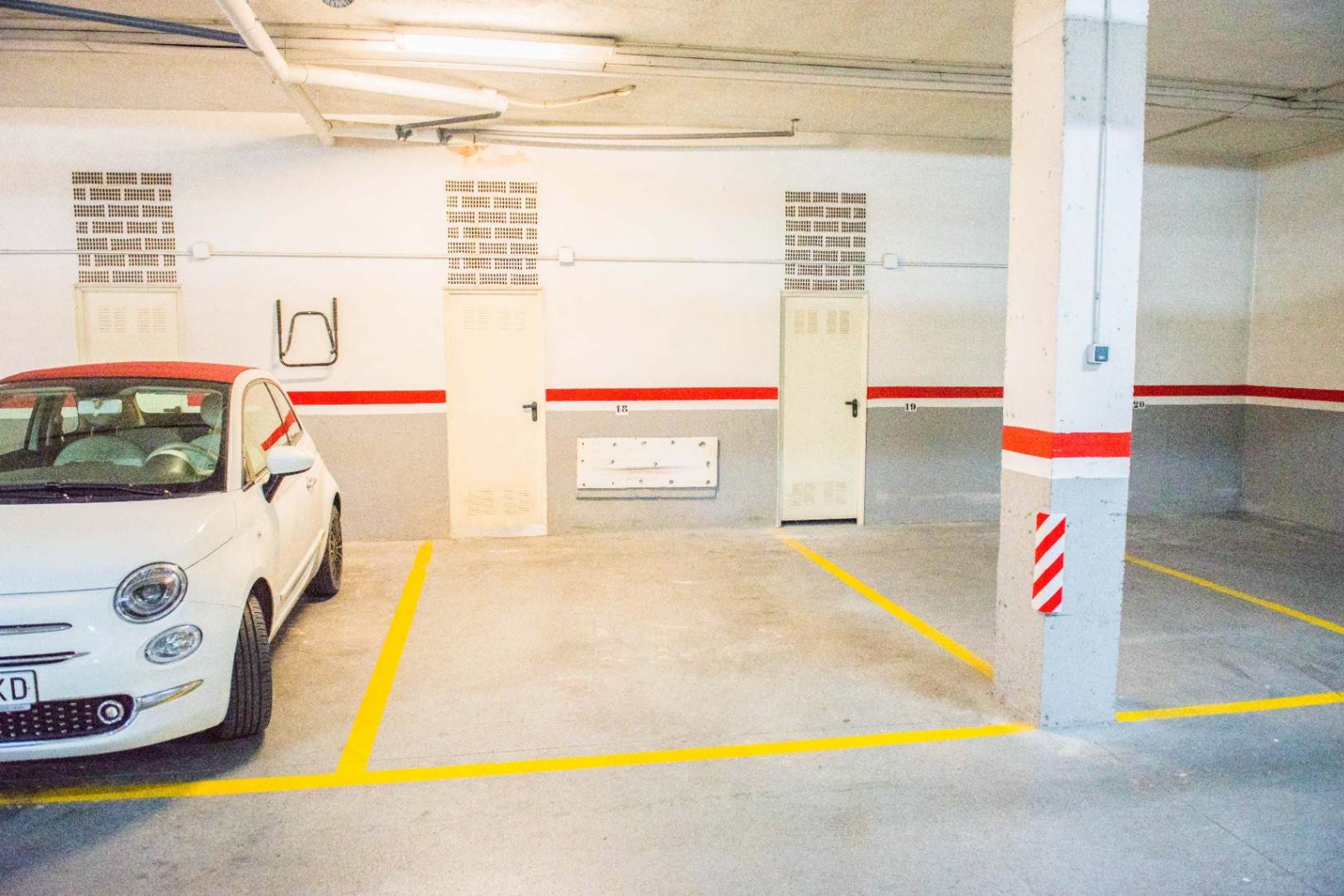 Emplacement de parking souterrain avec marquage au sol jaune, murs blancs et gris soulignés d'une bande rouge, et éclairage industriel au plafond.